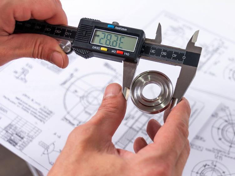 A quality engineer measuring a production part with calipers to ensure its compliance, with a reference diagram in the background.